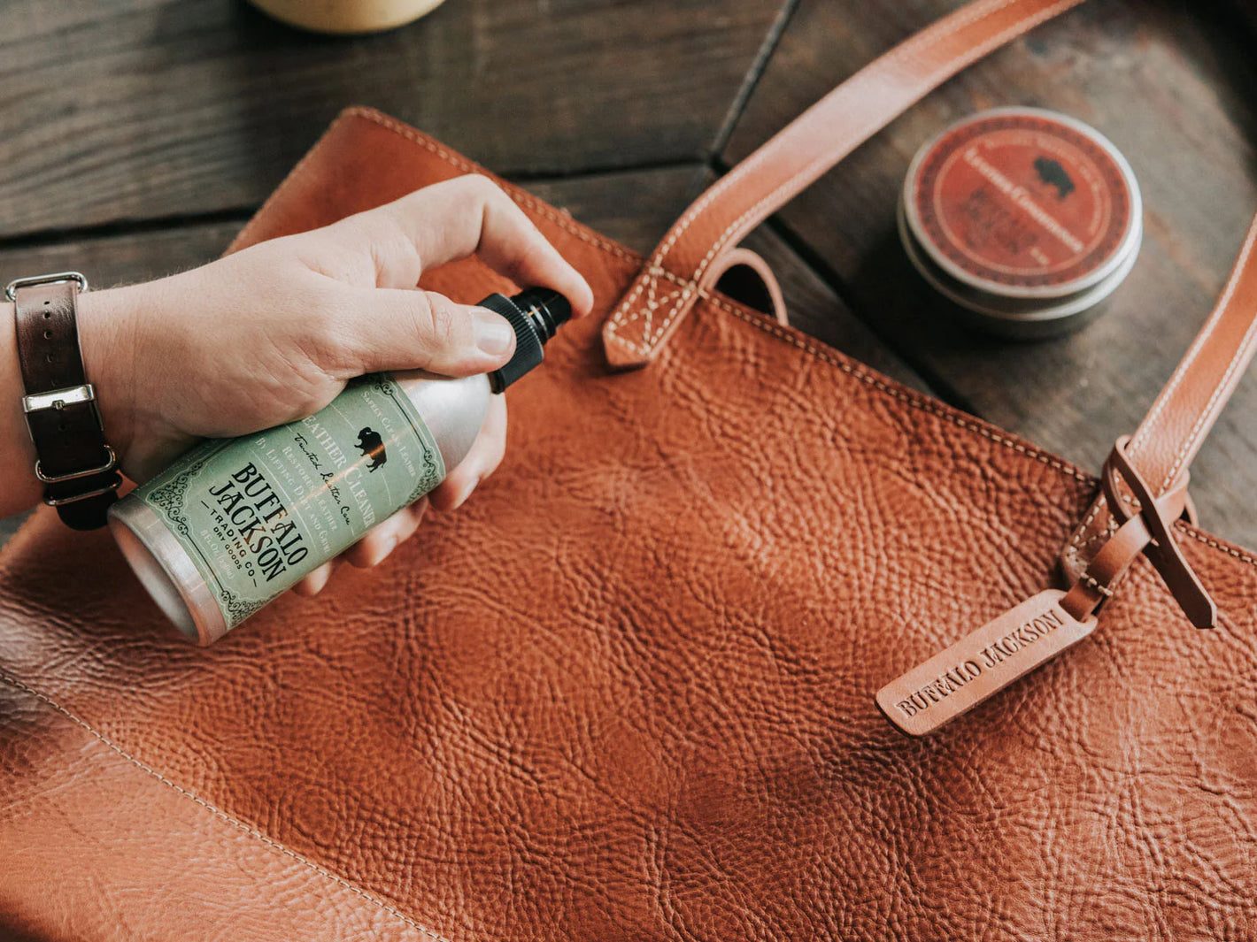 Person applying leather care product to a brown leather bag on a wooden surface.